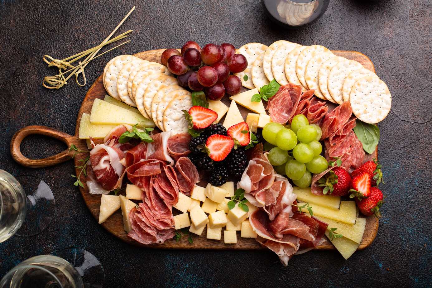 Charcuterie board filled with meats, cheeses and fruits with wine glasses next to it on a black counter