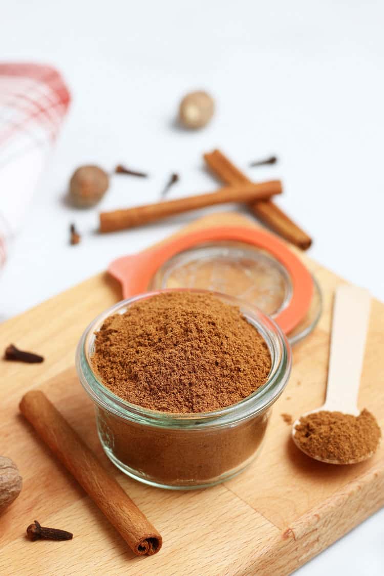 jar of pumpkin pie spice on a cutting board with a spoon next to it and a cinnamon stick and cloves scattered around