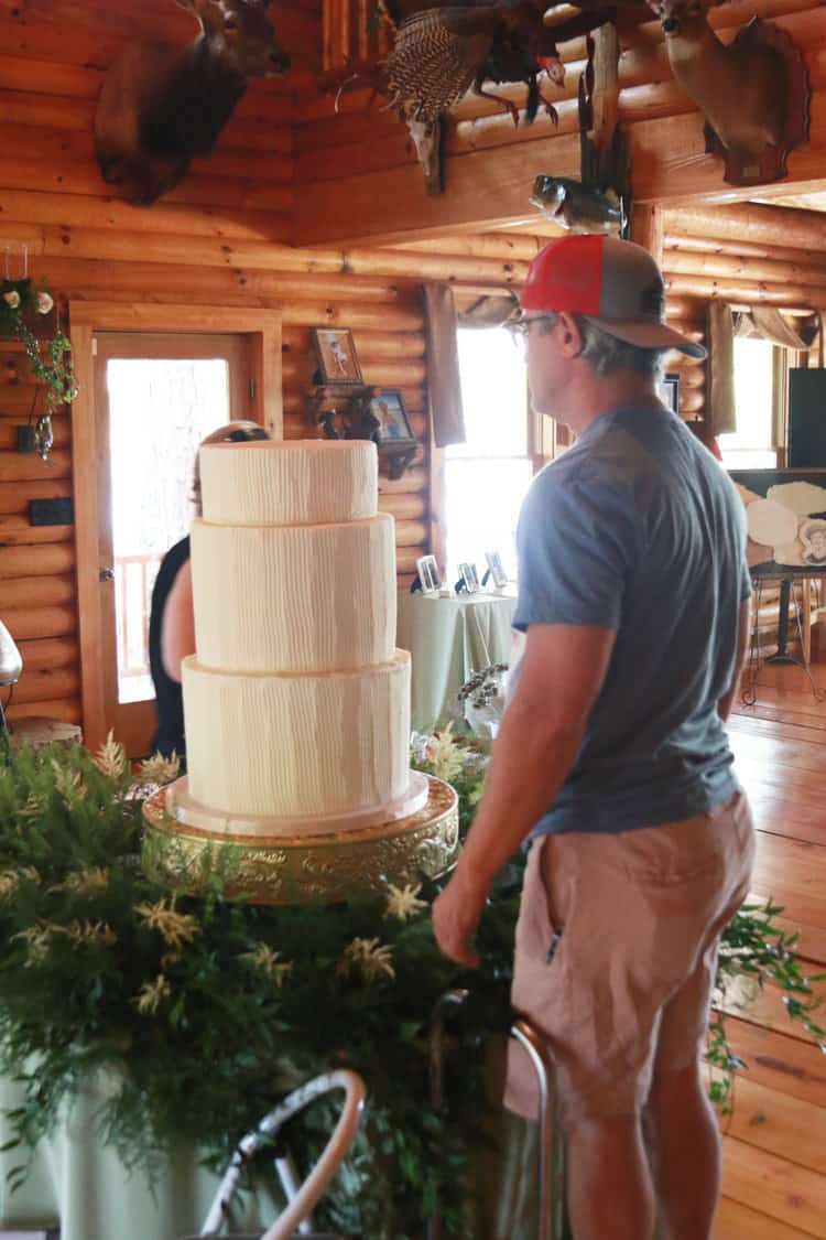 man looking at bottom three tiers of a wedding cake sitting on a cake stand and table filled with flowers